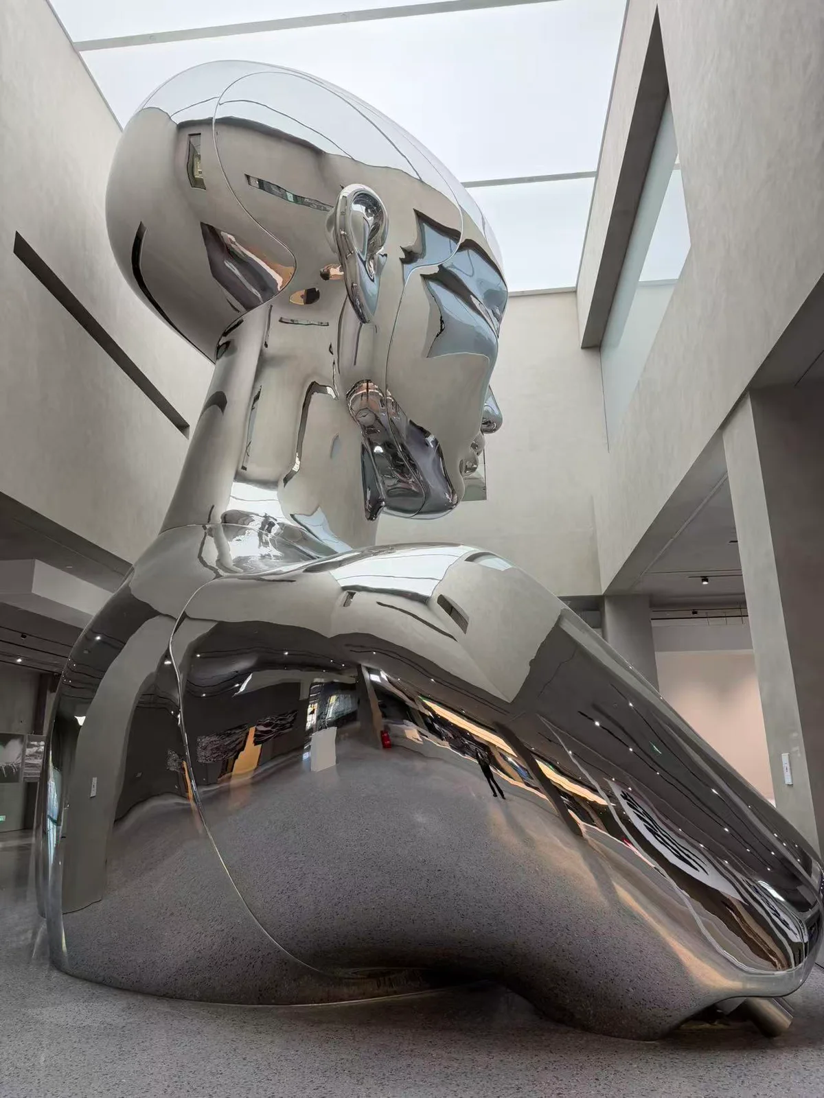 Full-height view of a large mirror stainless steel sculpture against skylight walls and lobby floor
