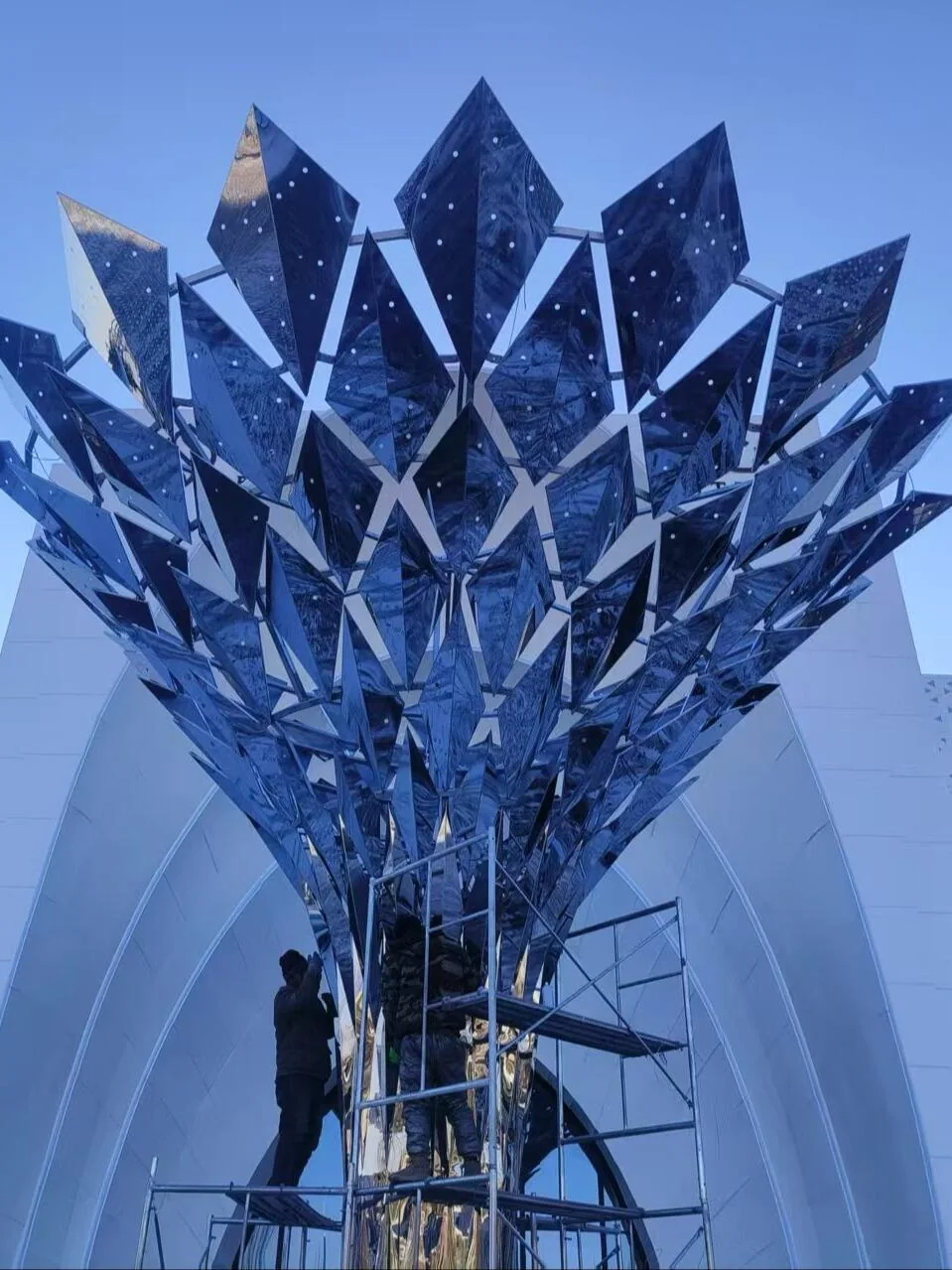 Reflective canopy sculpture in a construction setting with workers and access scaffold for scale reference
